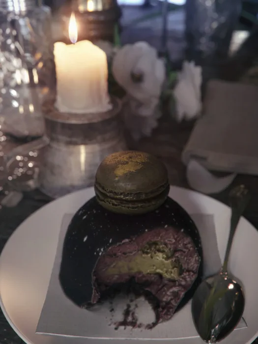 Wizualizacja pokazuje zbliżenie na drewniany blat stolika w restauracji, zbliżenie na talerz z połowicznie zjedzonym ciastkiem, z tyłu jest zapalona świeczka